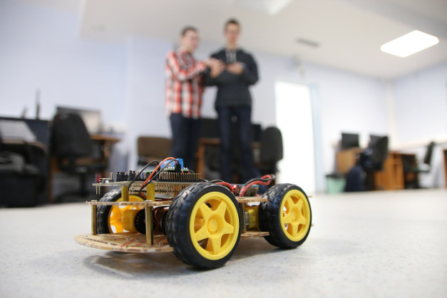 Zbudowany robot mobilny z żółtymi kołami i dwoma chłopcami w tle. Kontekst: warsztaty lub pracownia.