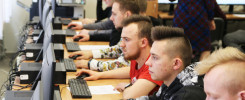 Uczniowie siedzą przy komputerach w sali lekcyjnej. Kluczowe obiekty: monitory, klawiatury, myszki, uczniowie.