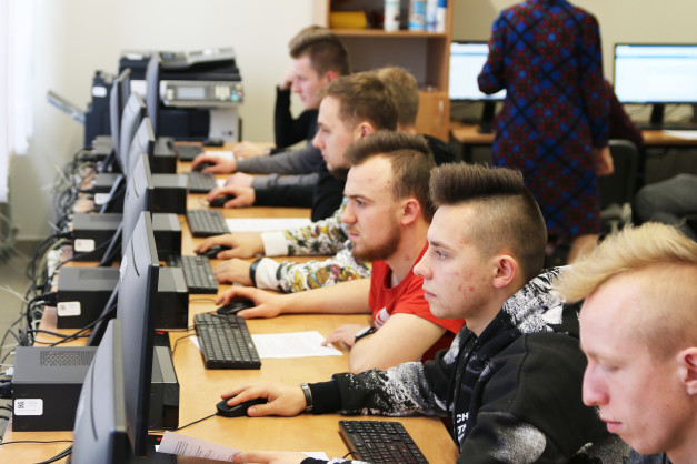 Uczniowie siedzą przy komputerach w sali lekcyjnej. Kluczowe obiekty: monitory, klawiatury, myszki, uczniowie.