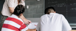 Nauczyciel pomaga uczniom w sali lekcyjnej. Kluczowe obiekty: nauczyciel, uczniowie, tablica, dokumenty. Kontekst: edukacja.