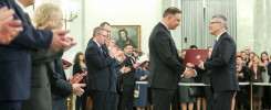 Prezydent RP wręcza dokument. W tle stoją ludzie, w tym mężczyzna z dyplomem.