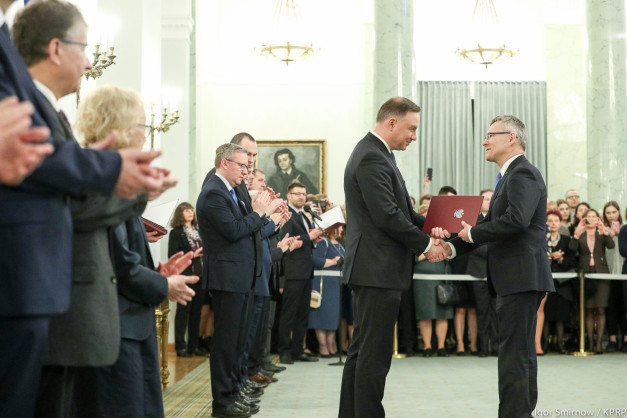 Prezydent RP wręcza dokument. W tle stoją ludzie, w tym mężczyzna z dyplomem.
