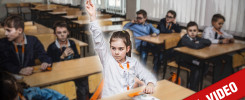 Dzieci na lekcji w sali z ławkami. Dziewczynka z podniesioną ręką. Kluczowe obiekty: ławki, dzieci, sala lekcyjna.