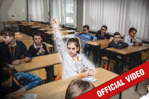 Dzieci na lekcji w sali z ławkami. Dziewczynka z podniesioną ręką. Kluczowe obiekty: ławki, dzieci, sala lekcyjna.