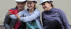 Trzy kobiety w białych kaskach robią selfie. Kobiety, kaski, telefon, kurtki.