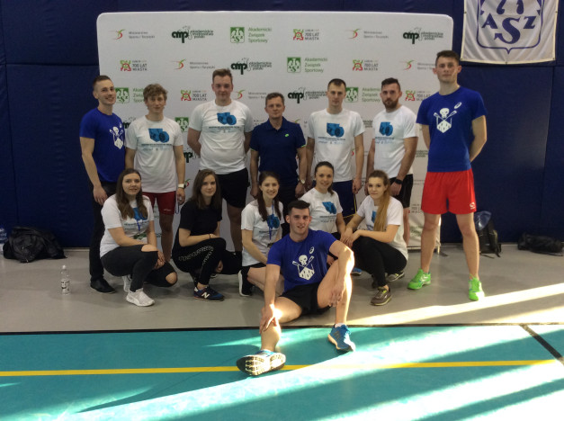 Grupa młodych ludzi w strojach sportowych pozuje do zdjęcia na tle baneru z logo.