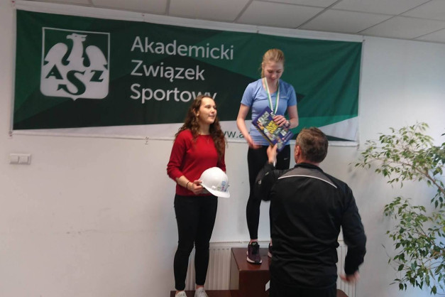Dwie kobiety na podium, jedna z medalem i książką, druga z kaskiem. Tło: baner "Akademicki Związek Sportowy".