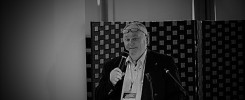 Mężczyzna w okularach przemawia do mikrofonu. Kontekst: konferencja, wykład. Obiekty: mężczyzna, mikrofon, okulary, podium.