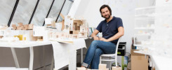 Mężczyzna siedzi przy biurku pełnym modeli architektonicznych. W tle duże okna i rysunki.