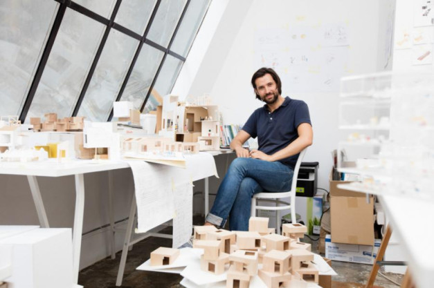 Mężczyzna siedzi przy biurku pełnym modeli architektonicznych. W tle duże okna i rysunki.