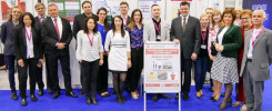 Grupa osób pozuje do zdjęcia z tablicą "Student-Inventor". Kluczowe obiekty: ludzie, tablica, plakaty.