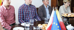 Cztery osoby siedzą przy stole. Na stole są flagi Polski i UE, ciastka, talerzyki i szklanki z wodą.