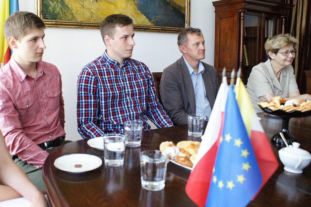 Cztery osoby siedzą przy stole. Na stole są flagi Polski i UE, ciastka, talerzyki i szklanki z wodą.