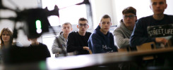 Grupa uczniów w klasie, przed nimi dron. Kluczowe obiekty: dron, uczniowie, klasa. Kontekst: lekcja, zajęcia praktyczne.