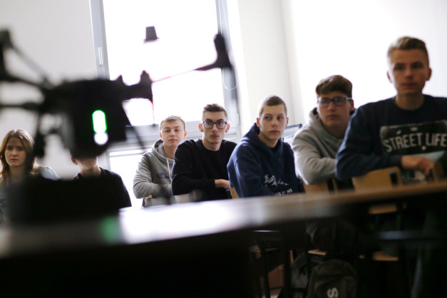 Grupa uczniów w klasie, przed nimi dron. Kluczowe obiekty: dron, uczniowie, klasa. Kontekst: lekcja, zajęcia praktyczne.