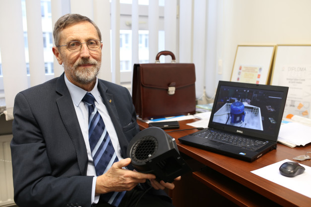 Starszy mężczyzna w garniturze trzyma wentylator. Na biurku laptop z filmem, teczka i klucze.