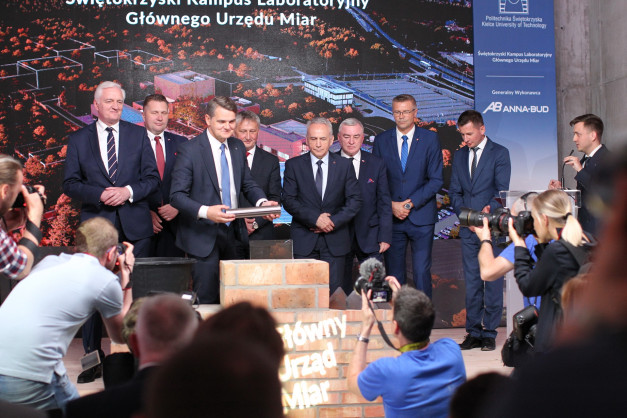 Mężczyźni w garniturach stoją przed ekranem z napisem "Świętokrzyski Kampus Laboratoryjny Głównego Urzędu Miar".