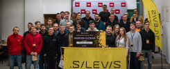 Grupa osób pozuje do zdjęcia z nagrodą główną Hackathonu Kielce 2023. Kluczowe obiekty: baner, ludzie, nagroda.