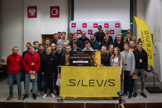 Grupa osób pozuje do zdjęcia z nagrodą główną Hackathonu Kielce 2023. Kluczowe obiekty: baner, ludzie, nagroda.