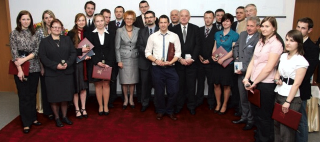 Grupa osób w strojach formalnych stoi na czerwonym dywanie, trzymając dyplomy.