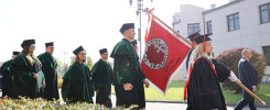 Osoby w togach akademickich i biretach idą z flagą. Kluczowe obiekty: togi, birety, flaga. Kontekst: uroczystość akademicka.
