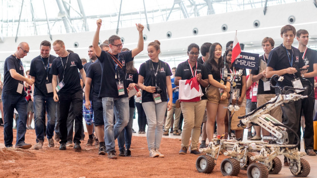 Grupa młodych ludzi w czarnych koszulkach z identyfikatorami, z flagą Polski i robotem. Konkurs robotyki.