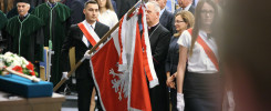 Mężczyzna w garniturze niesie polską flagę. W tle stoją ludzie w togach i strojach galowych. Kontekst: inauguracja roku akademickiego.