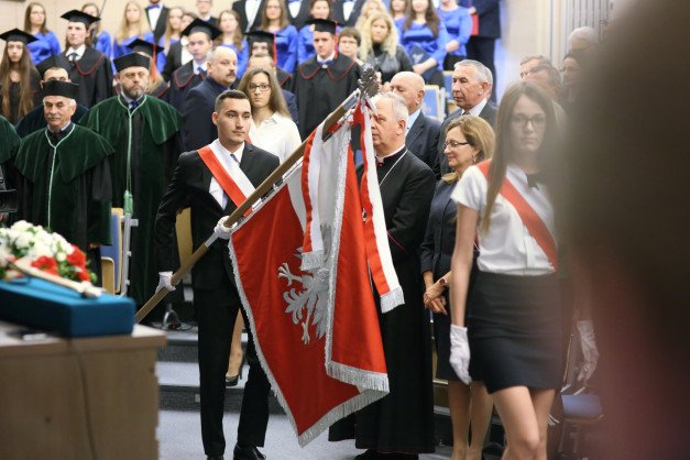 Mężczyzna w garniturze niesie polską flagę. W tle stoją ludzie w togach i strojach galowych. Kontekst: inauguracja roku akademickiego.
