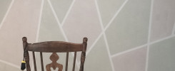 Drewniane krzesło z ozdobnym oparciem w kształcie serca wisi na nim mały przedmiot. Tło to ściana z geometrycznym wzorem.