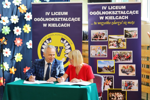 Mężczyzna i kobieta siedzą przy stole, podpisując dokumenty. W tle banery z logo IV Liceum Ogólnokształcącego w Kielcach.
