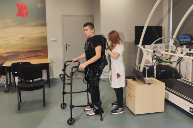 Chłopiec w egzoszkielecie rehabilitacyjnym stoi przy chodziku. Obok niego stoi kobieta w białej bluzie.
