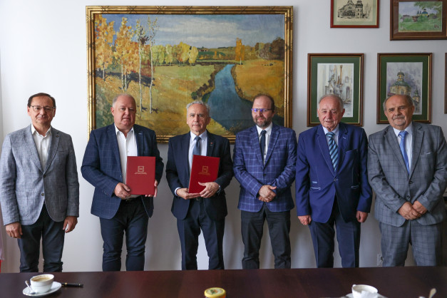 Sześciu mężczyzn w garniturach stoi przed obrazem. Trzech z nich trzyma czerwone teczki.
