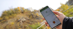 Dłoń trzyma smartfon z aplikacją Geopark Kielce. W tle widać wzgórze z drzewami.