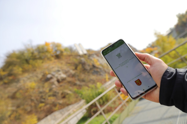 Dłoń trzyma smartfon z aplikacją Geopark Kielce. W tle widać wzgórze z drzewami.