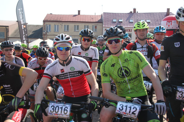 Kolarze w strojach sportowych i kaskach stoją przed budynkami. Widoczne rowery, flagi i numery startowe.