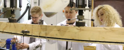 Trzy osoby w białych fartuchach pracują przy drewnianej belce z aparaturą. Kontekst: laboratorium, testowanie materiałów.