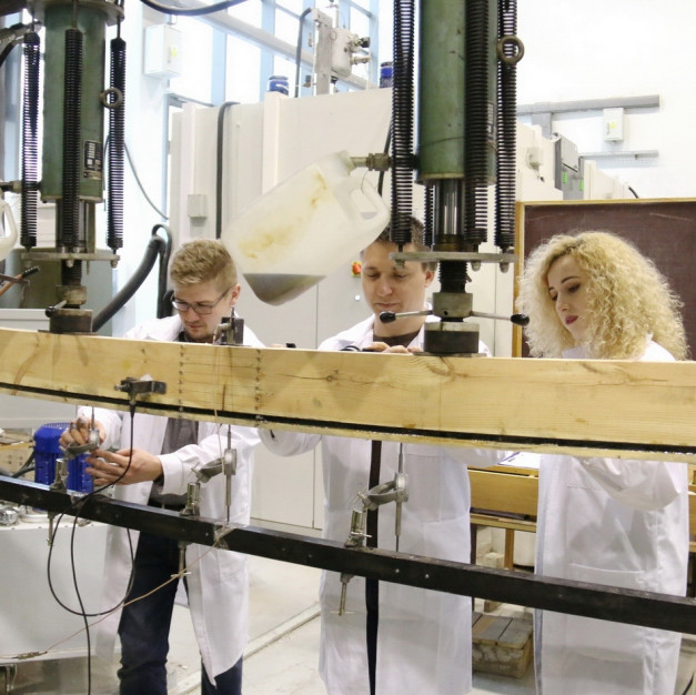 Trzy osoby w białych fartuchach pracują przy drewnianej belce z aparaturą. Kontekst: laboratorium, testowanie materiałów.
