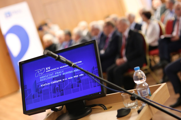 Monitor z napisem "XV Konferencja Naukowo-Techniczna", mikrofon, butelka wody. Kontekst: wydarzenie naukowe.