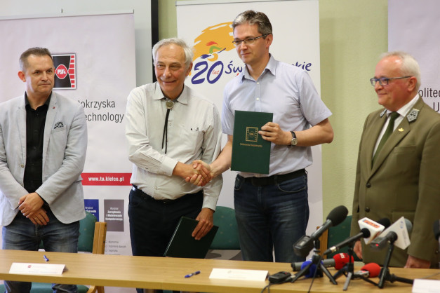 Czterech mężczyzn stoi przy stole. Dwóch z nich ściska sobie dłonie, jeden trzyma zieloną teczkę.