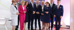 Grupa osób w eleganckich strojach stoi przed banerem z napisem "Budowa Kampusu". Kluczowe obiekty: ludzie, baner.