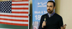 Mężczyzna z mikrofonem przemawia obok flagi USA i banera "camp leaders Work & Travel".