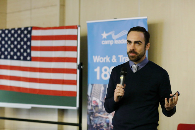 Mężczyzna z mikrofonem przemawia obok flagi USA i banera "camp leaders Work & Travel".