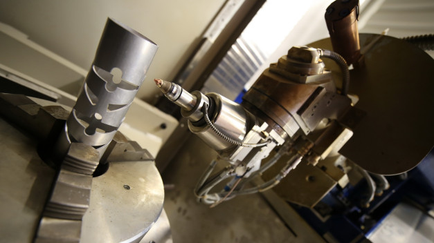 Metalowy element z wycięciami w uchwycie maszyny CNC. W tle widoczna głowica robocza.