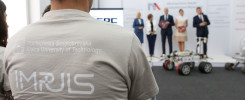 Osoba w koszulce z logo Politechniki Świętokrzyskiej. W tle panel z logo, osoby i robot.