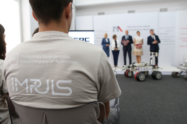 Osoba w koszulce z logo Politechniki Świętokrzyskiej. W tle panel z logo, osoby i robot.
