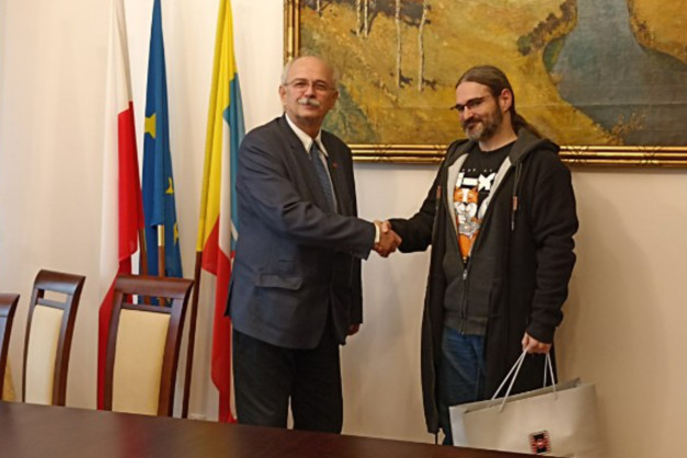 Dwóch mężczyzn wita się na tle flag i obrazu. Kluczowe obiekty: mężczyźni, flagi, obraz, krzesła. Kontekst: spotkanie.