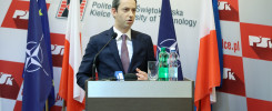 Mężczyzna w garniturze przemawia za mównicą. W tle flagi NATO, Polski i UE. Kontekst: konferencja prasowa.
