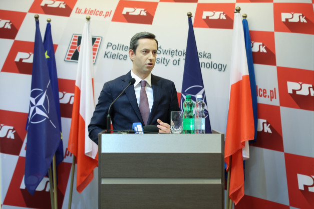 Mężczyzna w garniturze przemawia za mównicą. W tle flagi NATO, Polski i UE. Kontekst: konferencja prasowa.