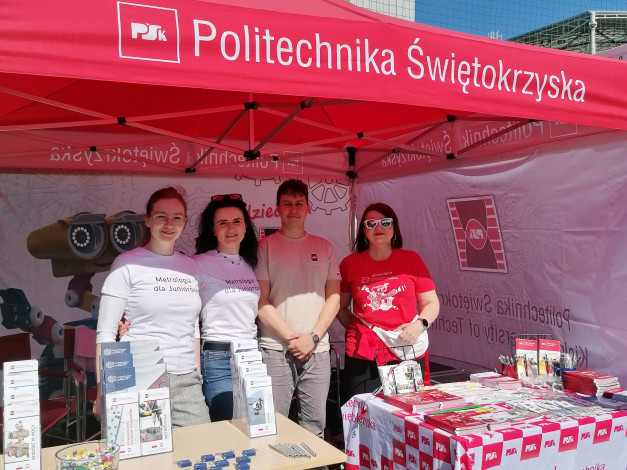 Cztery osoby stoją pod czerwonym namiotem z napisem "Politechnika Świętokrzyska". Na stole leżą materiały promocyjne.