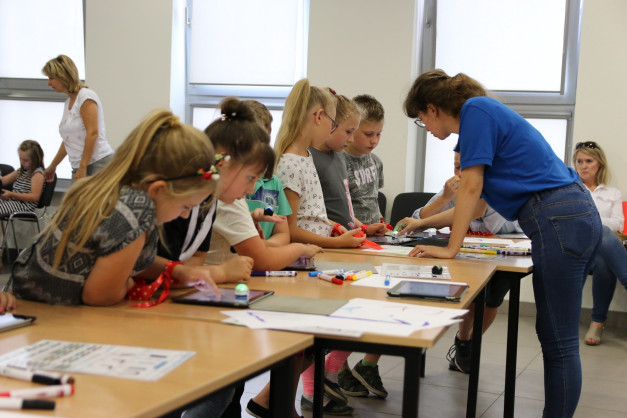 Dzieci i dorośli przy stołach z tabletami i robotami. Kontekst: warsztaty, nauka, technologia.